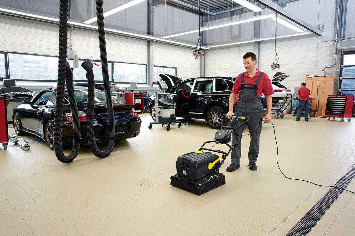 A man in a red shirt and gray overalls operates a Karcher BR 40/10C black and yellow floor buffer machine in a bright auto repair garage, with cars and service equipment visible in the background.