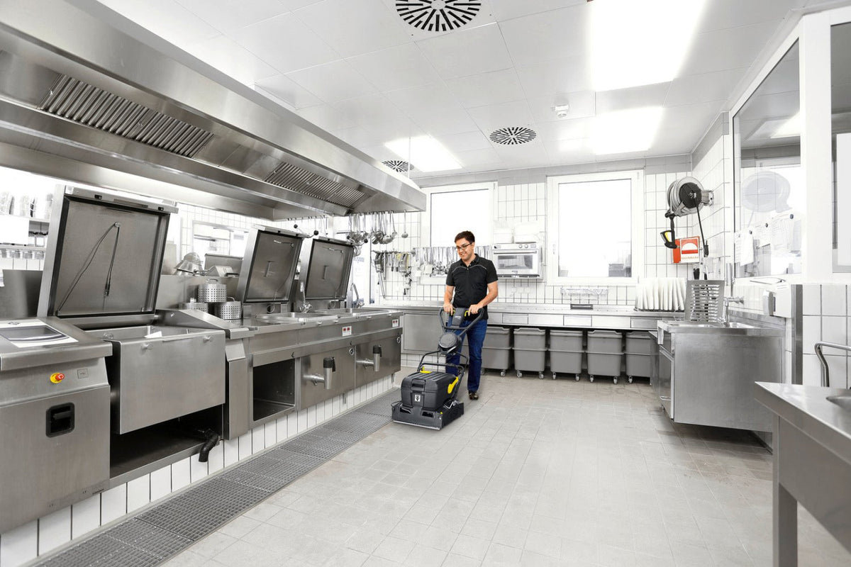 A commercial kitchen with stainless steel counters and equipment, featuring a man operating a Karcher BR 40/10C floor scrubber on the light gray tiled floor.